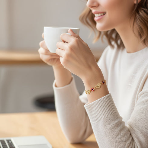 A woman smiles holding a white mug, wearing a cream sweater and a Heart Rolo Link bracelet, seated at a wooden table with her laptop.